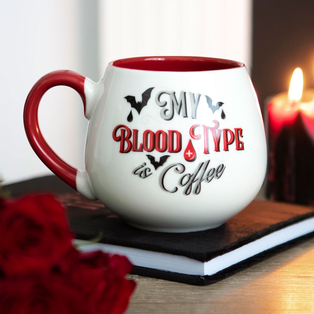 White rounded ceramic mug with red handle and interior, printed with My Blood Type is Coffee slogan with bat and blood drop details, styled with a candle and red roses