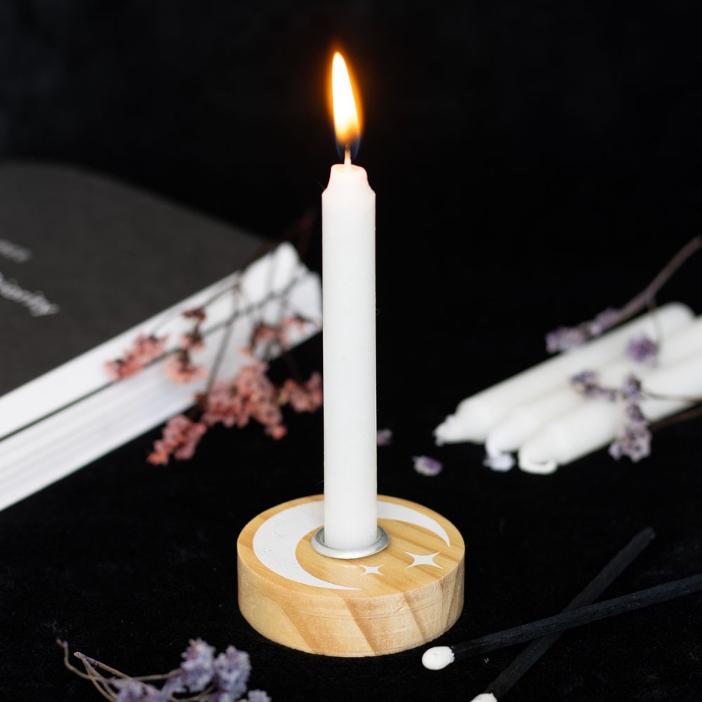 Round natural wood spell candle holder with white crescent moon and stars design, with a lit white taper candle in the metal insert, styled on a dark surface with dried flowers and an open book