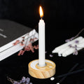Round natural wood spell candle holder with white crescent moon and stars design, with a lit white taper candle in the metal insert, styled on a dark surface with dried flowers and an open book