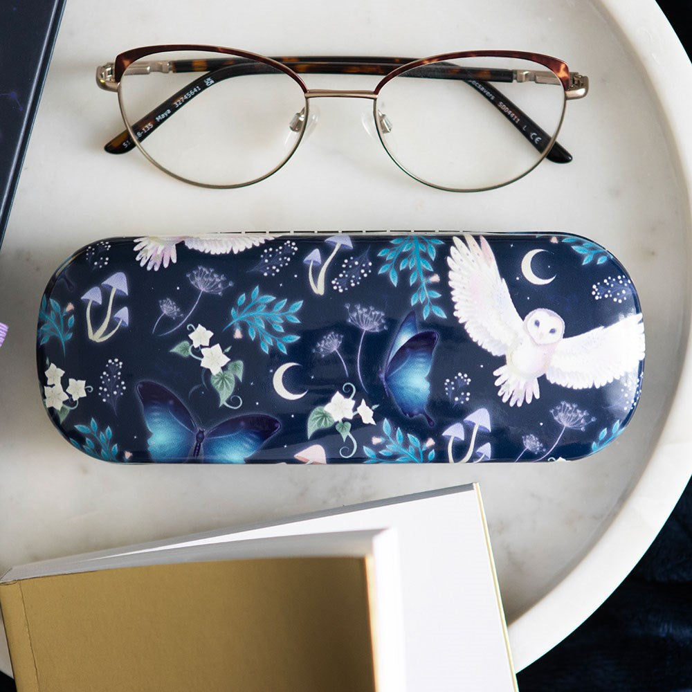 Navy hard shell glasses case with a magical night design featuring a white barn owl in flight, blue morpho butterflies, crescent moons, mushrooms, and botanical leaves, styled on a marble tray with tortoiseshell glasses and a book