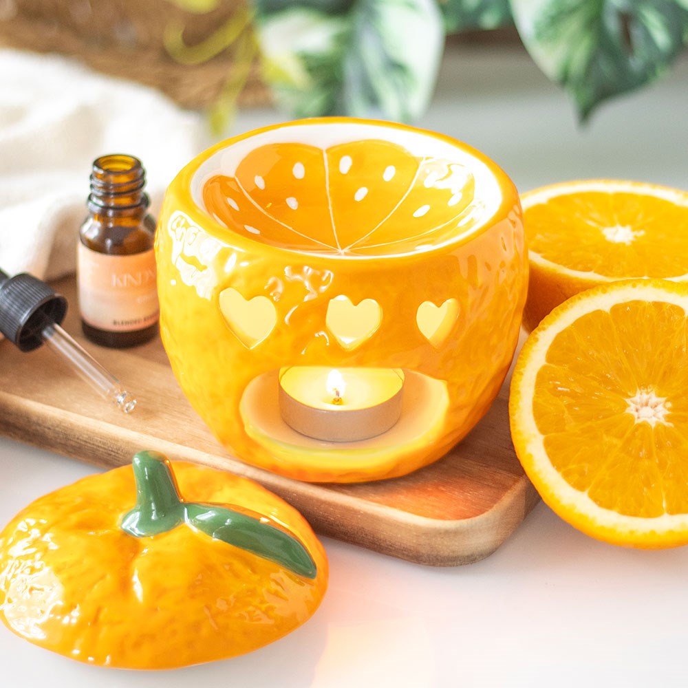 Orange fruit shaped ceramic oil burner with heart cut-outs and lit tealight, styled on a wooden board with fresh orange slices and an essential oil bottle