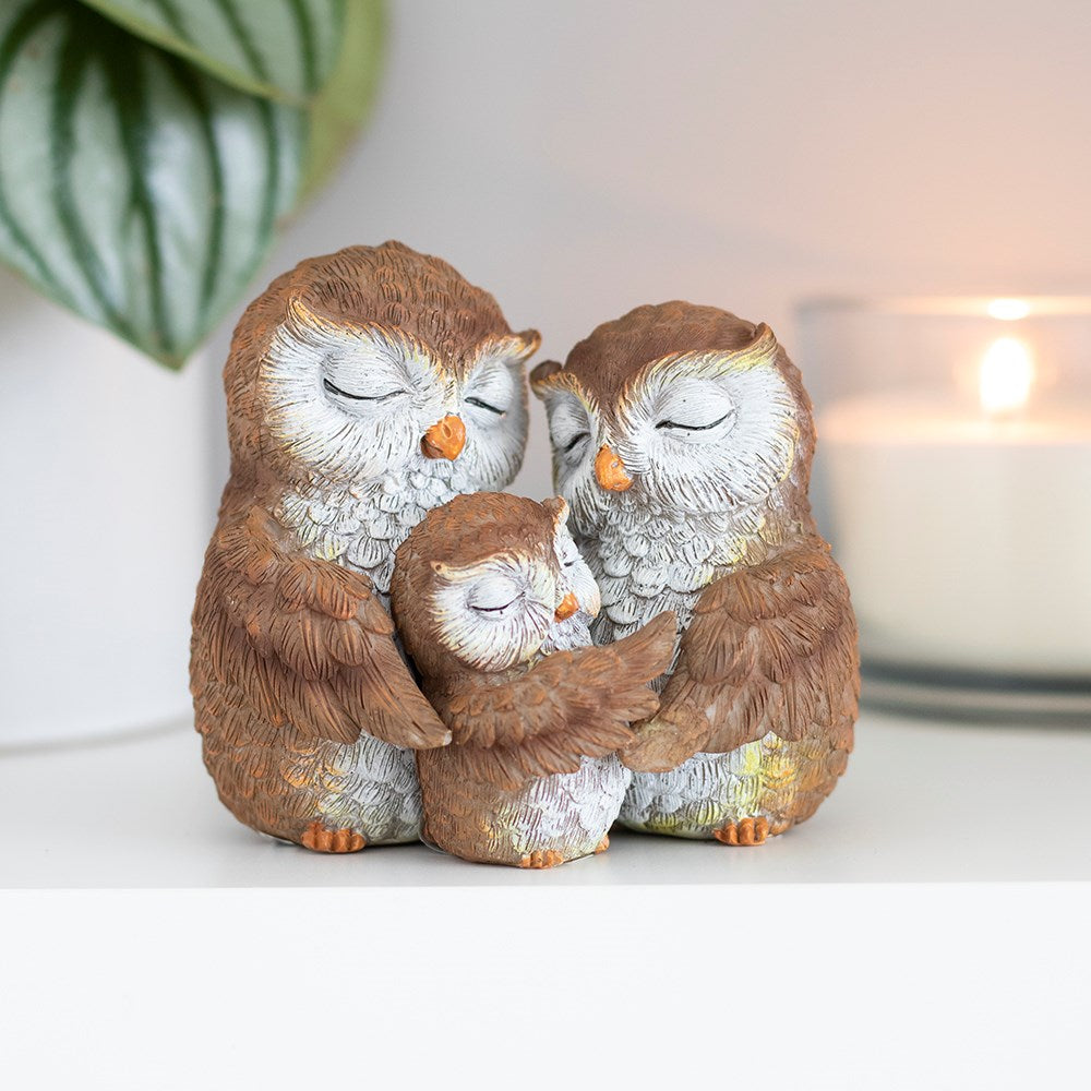 Resin owl family ornament showing two adult owls and a small owlet huddled together with eyes closed, in warm brown and grey tones, styled with a candle and green plant in the background