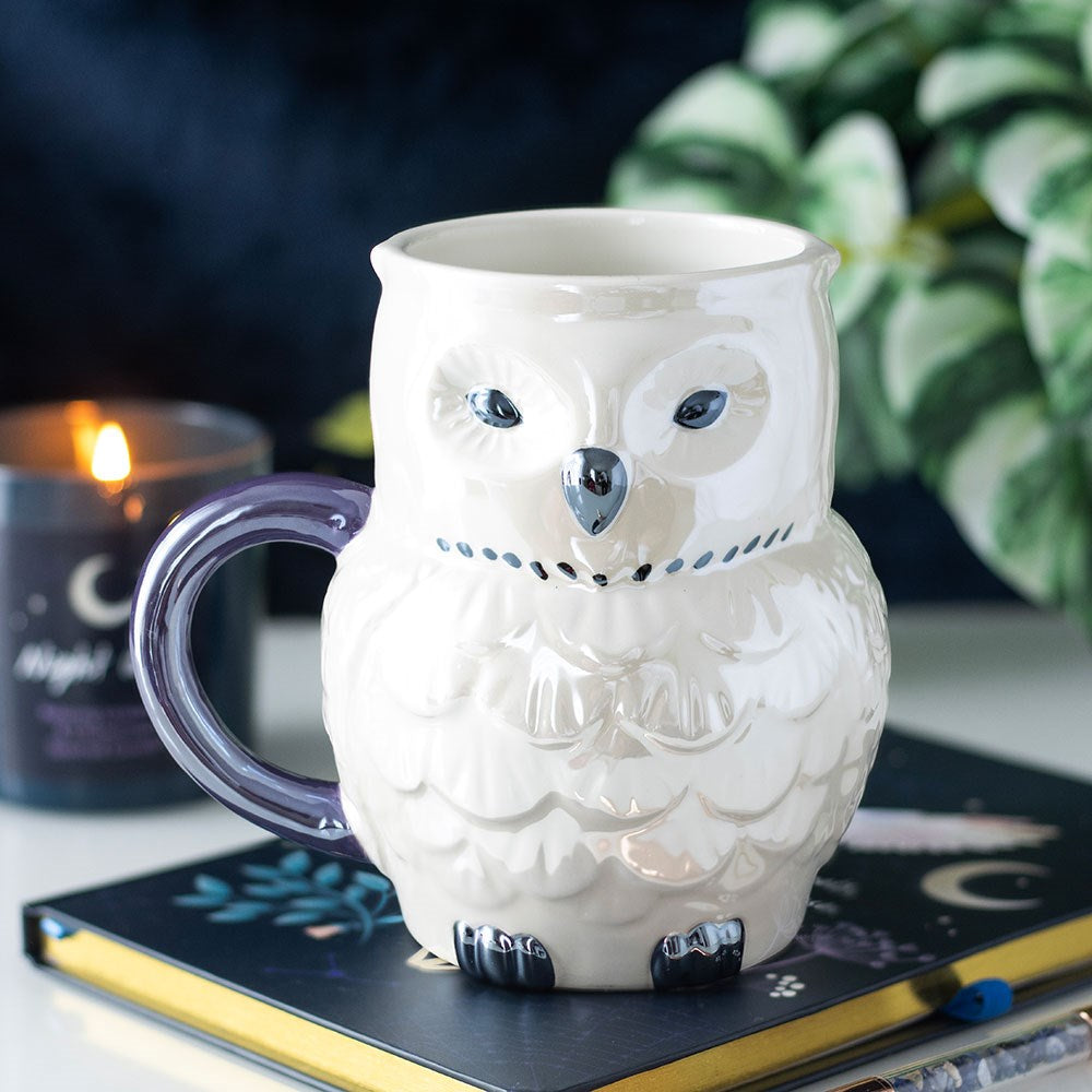 White ceramic owl shaped mug with embossed feather detailing, silver beak and talons, and a grey crescent moon handle, styled on a dark notebook next to a lit candle and green plant