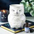 White iridescent ceramic owl shaped oil burner with silver beak and eyes, styled on a journal with a candle and essential oil bottle