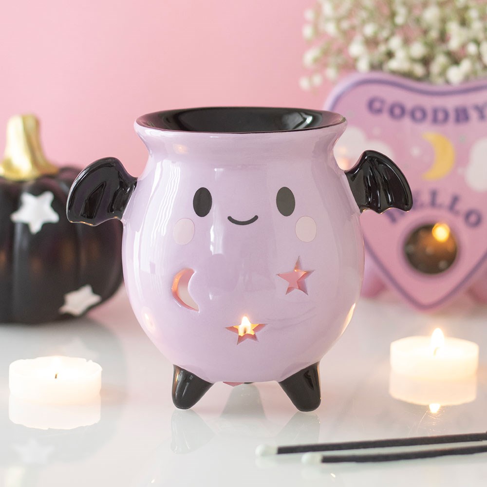 Pastel pink ceramic cauldron oil burner with black bat wings, smiley face, and star and moon cut-outs, glowing with a lit tealight, styled with candles and a ouija board decoration