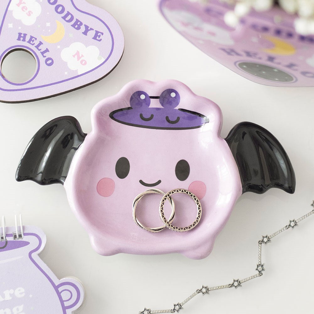 Pastel purple ceramic bat trinket dish with black wings and kawaii smiley face, holding two silver rings, styled with ouija board and star chain necklace