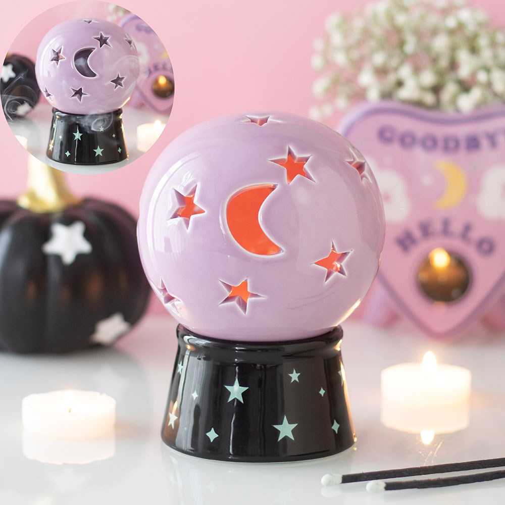 Pastel purple ceramic crystal ball tealight holder with star and moon cut-outs glowing orange, on a black star-print base, styled with candles and a ouija board decoration