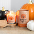 Pumpkin spice fragranced candle in a glass jar with cork lid and jack-o-lantern label lit and glowing, beside its cylindrical kraft gift tube, surrounded by decorative pumpkins on a wooden surface