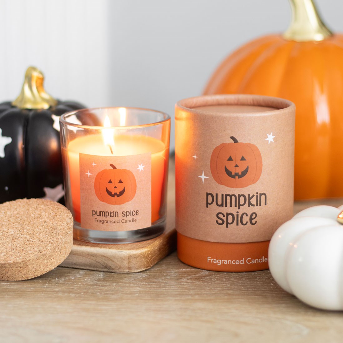 Pumpkin spice fragranced candle in a glass jar with cork lid and jack-o-lantern label lit and glowing, beside its cylindrical kraft gift tube, surrounded by decorative pumpkins on a wooden surface