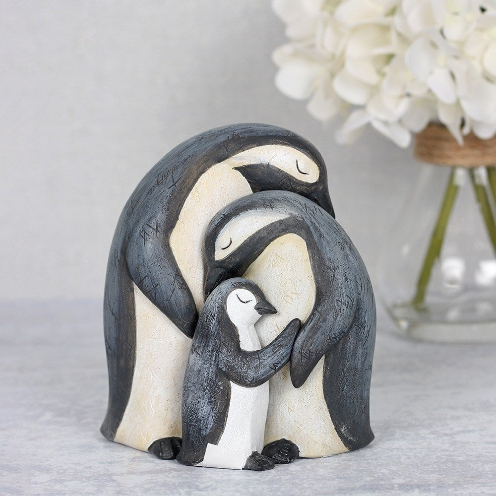 Resin penguin family ornament showing two adult penguins and a chick hugging, with a rustic grey and cream textured finish, styled with white hydrangeas in the background
