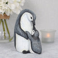 Resin penguin parent and child ornament, an adult penguin wrapping its flipper around a small chick, with a rustic grey and white textured finish, styled with white hydrangeas and a candle in the background