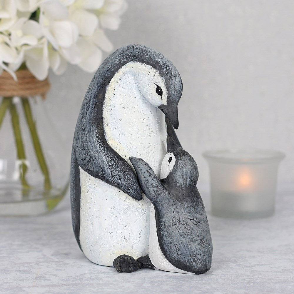 Resin penguin parent and child ornament, an adult penguin wrapping its flipper around a small chick, with a rustic grey and white textured finish, styled with white hydrangeas and a candle in the background