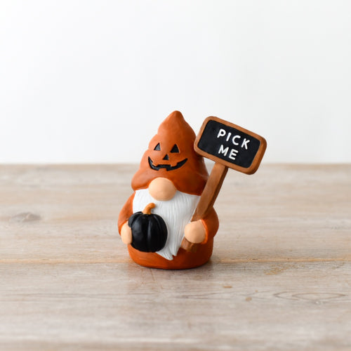 Orange ceramic autumn gonk figurine with jack-o'-lantern hat, holding a black pumpkin and a "Pick Me" chalkboard sign, on a wooden surface

