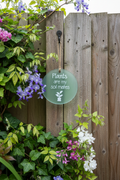 Round frosted glass "Plants Are My Soil Mates" hanging sign with twine, displayed on a rustic wooden garden fence surrounded by wisteria and flowers