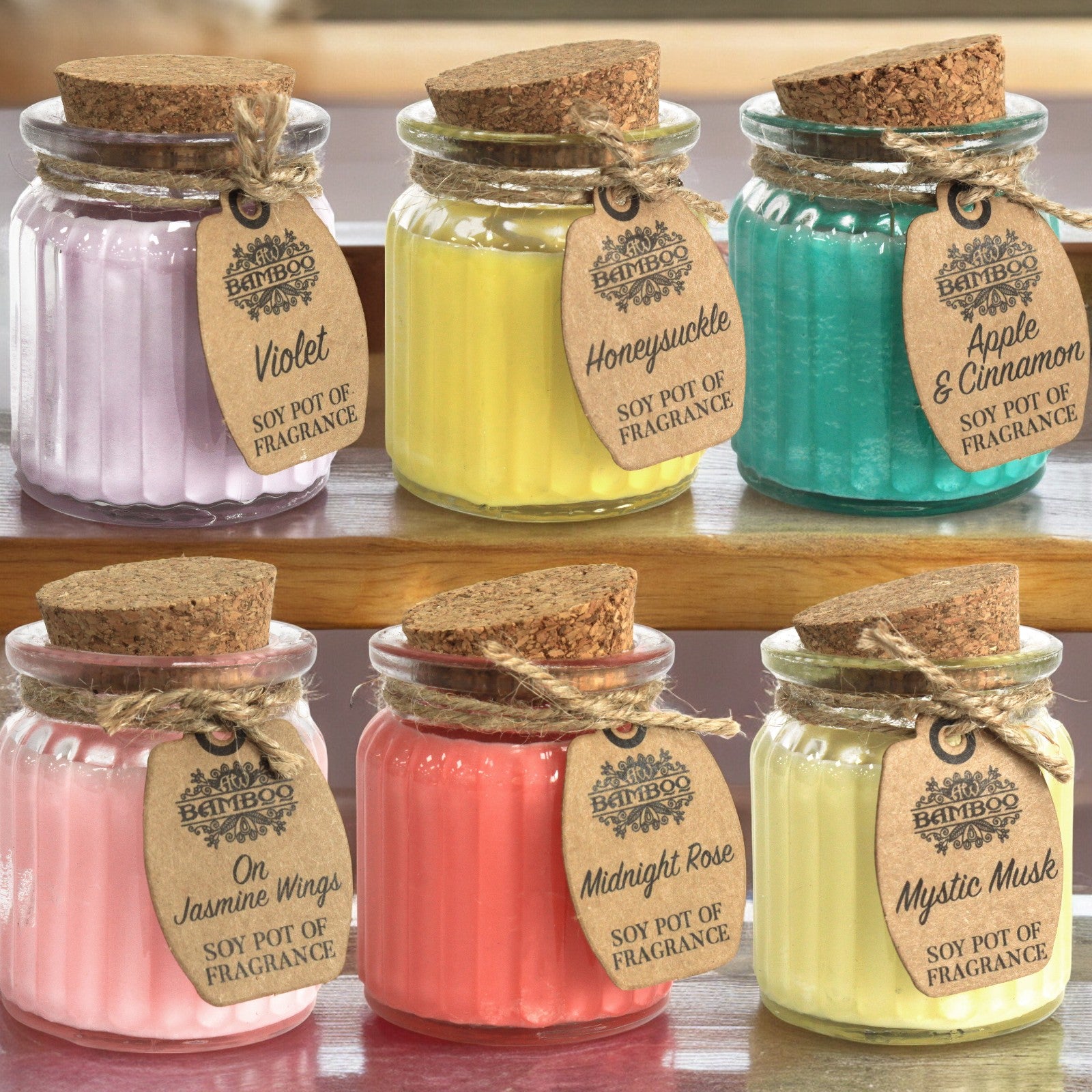 Six colourful soy wax fragrance jar candles in ribbed glass pots with cork lids and kraft paper tags, displayed on a wooden shelf