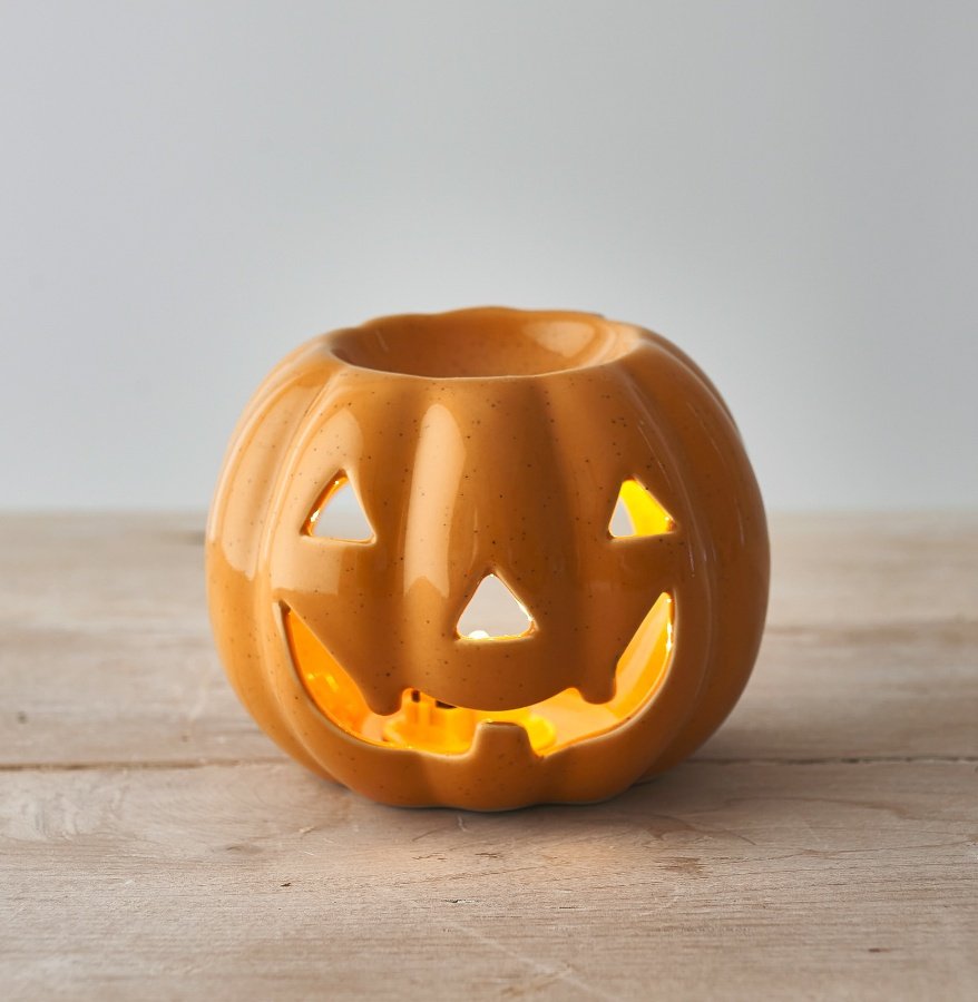 Orange ceramic pumpkin jack-o'-lantern oil burner with glowing cut-out face, sitting on a pale wooden surface with warm candlelight glow