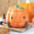 Cute smiling orange ceramic pumpkin oil burner and wax warmer with little brown legs and green stem lid, styled on a wooden board with autumn leaves and essential oil bottle