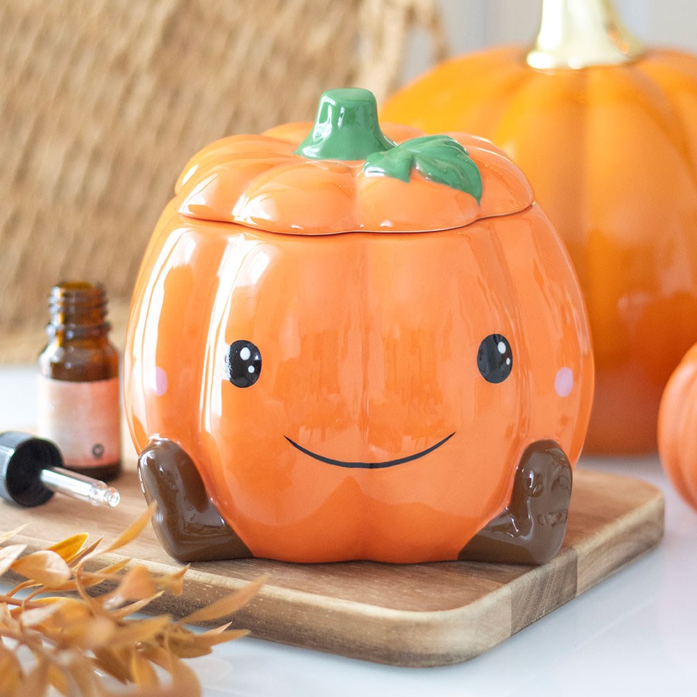 Cute smiling orange ceramic pumpkin oil burner and wax warmer with little brown legs and green stem lid, styled on a wooden board with autumn leaves and essential oil bottle