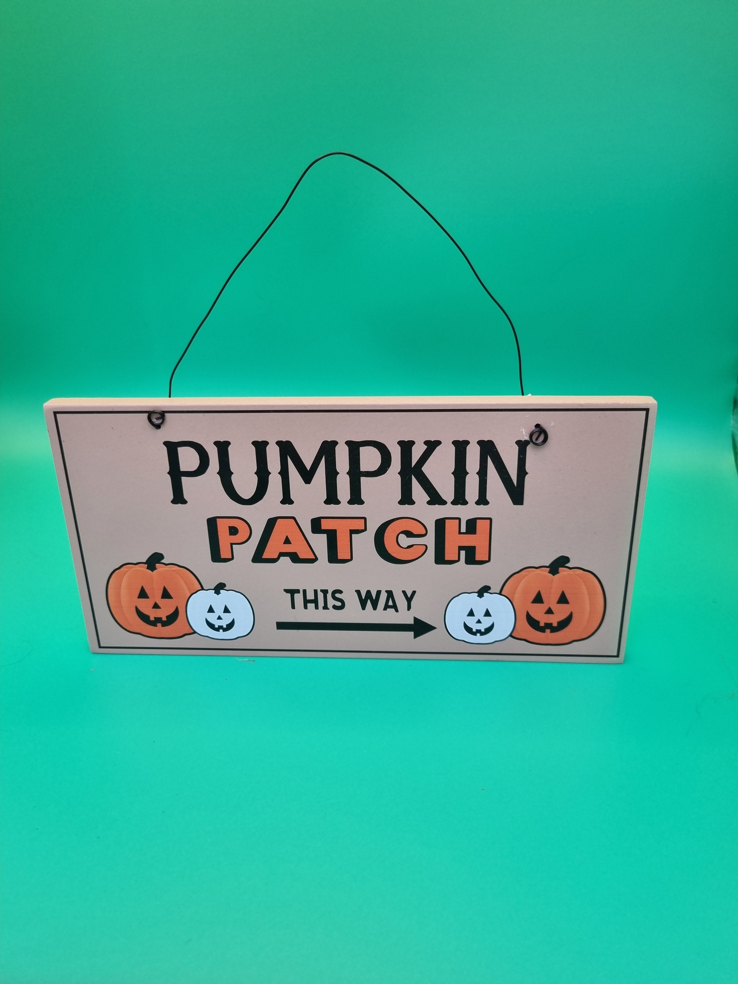 Wooden hanging sign reading Pumpkin Patch This Way with orange and white jack-o'-lantern illustrations and a directional arrow, hung with black wire