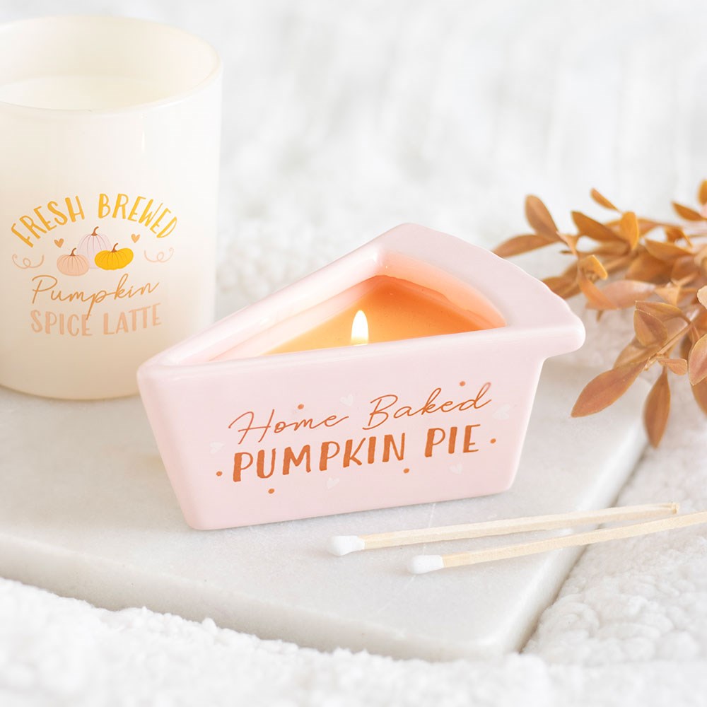 Pink pie slice shaped pumpkin pie scented candle with lit flame, styled on a white tray with autumn foliage and a pumpkin spice latte candle