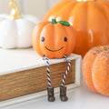 Cute smiling orange resin pumpkin shelf sitter decoration with twisted rope legs and brown boots perched on a wooden ledge surrounded by decorative pumpkins