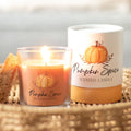 Lit pumpkin spice scented candle in an orange glass jar with pumpkin label, displayed alongside its white and orange gift box on a woven basket tray