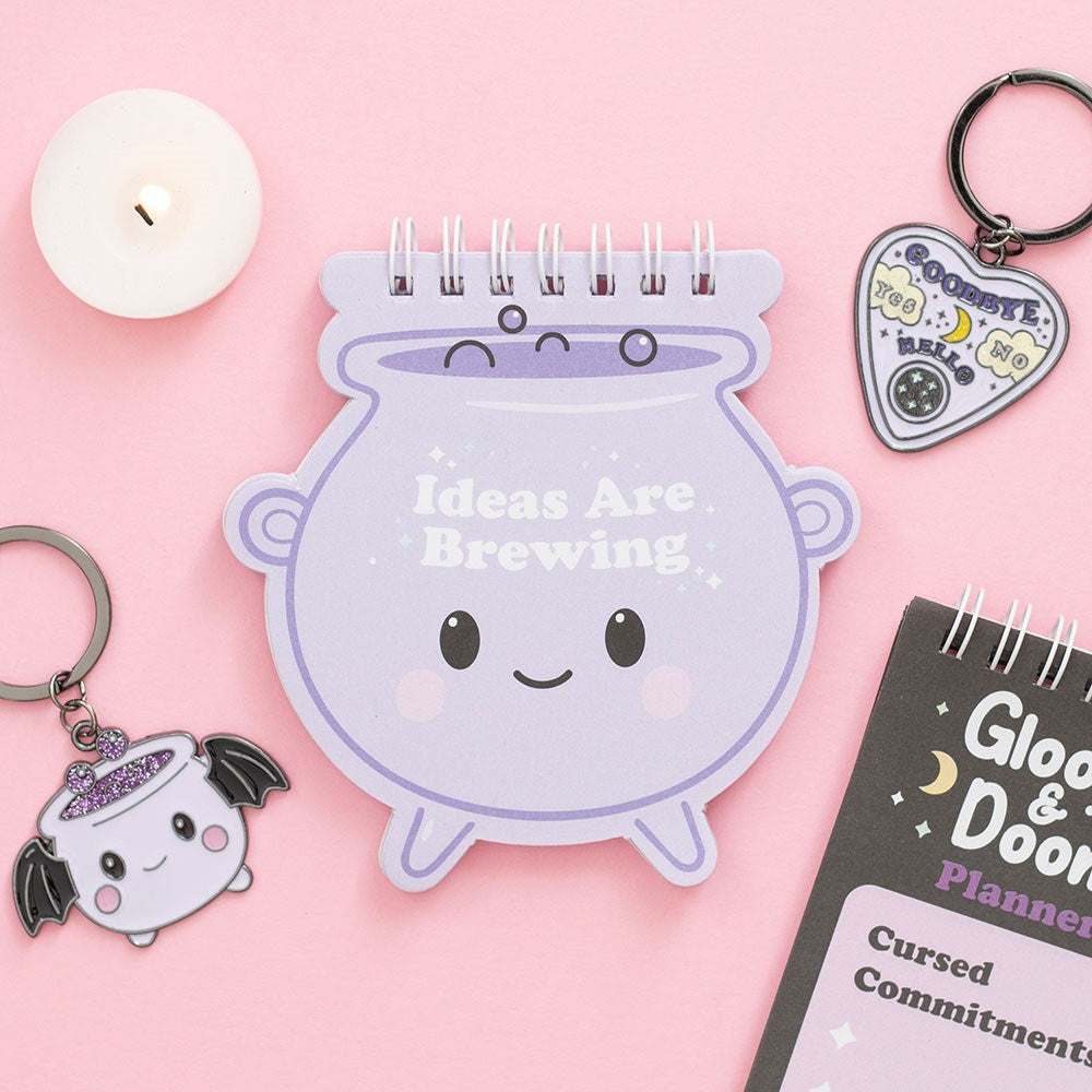 Mini pastel purple cauldron shaped notepad with Ideas Are Brewing design, styled on a pink background with keyrings, a candle and the Gloom & Doom planner
