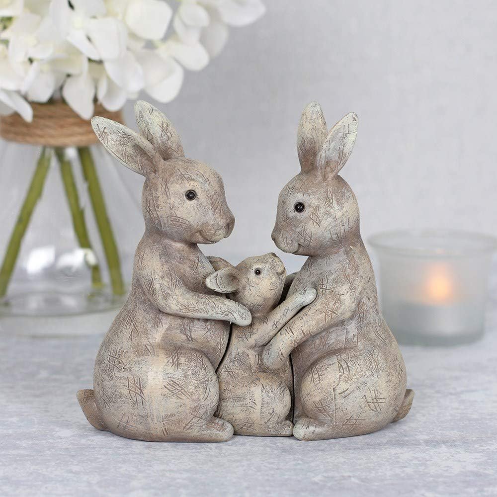 Resin bunny rabbit family ornament showing two adult rabbits cradling a baby rabbit, with a rustic grey textured finish, styled with white hydrangeas and a candle in the background