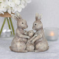Resin bunny rabbit family ornament showing two adult rabbits cradling a baby rabbit, with a rustic grey textured finish, styled with white hydrangeas and a candle in the background