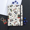 Retro 80s design A5 hardback lined notebook with cassette tape, VHS and arcade game pattern, styled with blue dice and a matching keyring on a dark surface