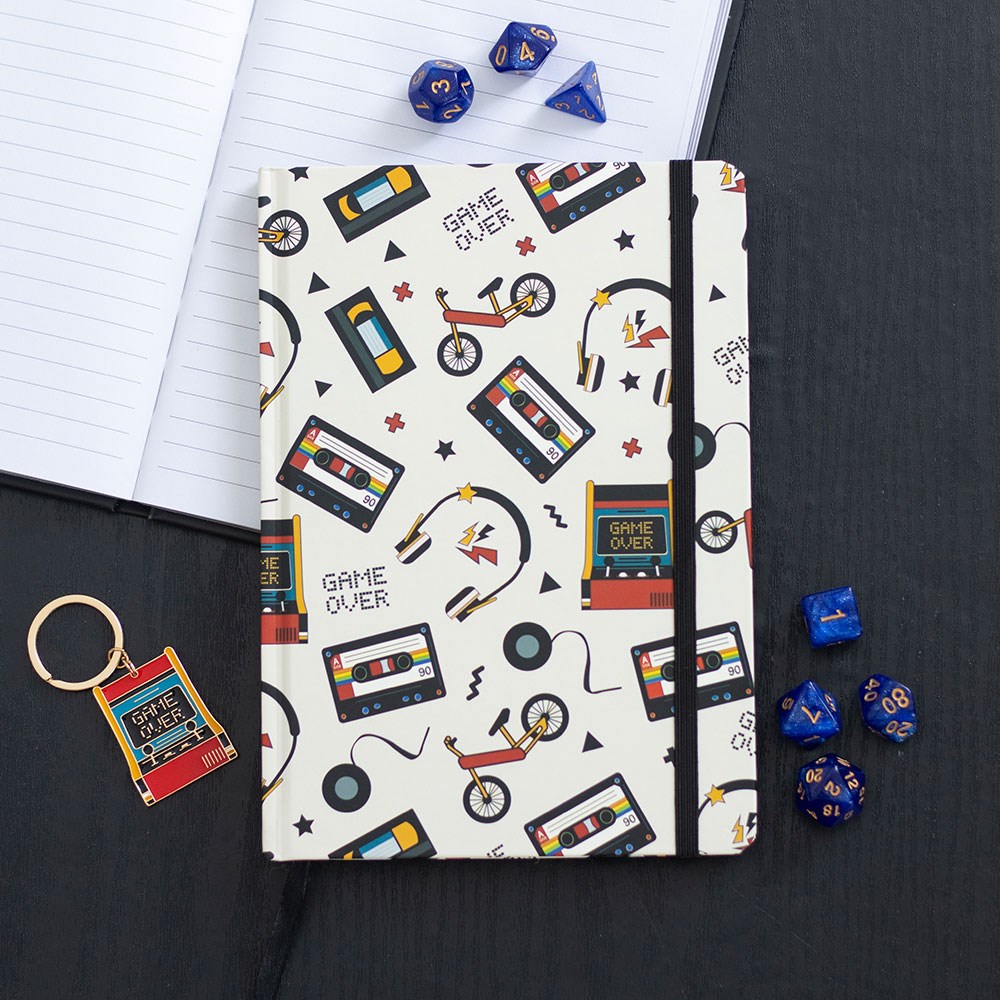 Retro 80s design A5 hardback lined notebook with cassette tape, VHS and arcade game pattern, styled with blue dice and a matching keyring on a dark surface