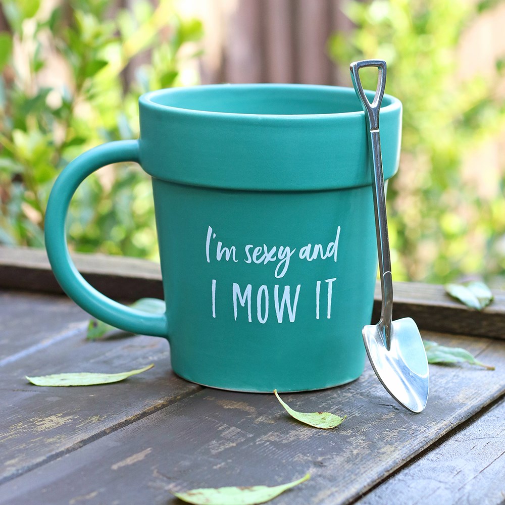 Teal ceramic plant pot shaped mug with "I'm sexy and I MOW IT" text in white, displayed outdoors on a wooden surface with a stainless steel shovel spoon leaning against it and scattered leaves