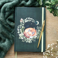 Dark green hardback A5 lined notebook with a sleeping fox curled in a botanical leaf wreath with a crescent moon, stars and a small bird on the cover, displayed on a wooden surface with a gold pen, green blanket and white gypsophila flowers