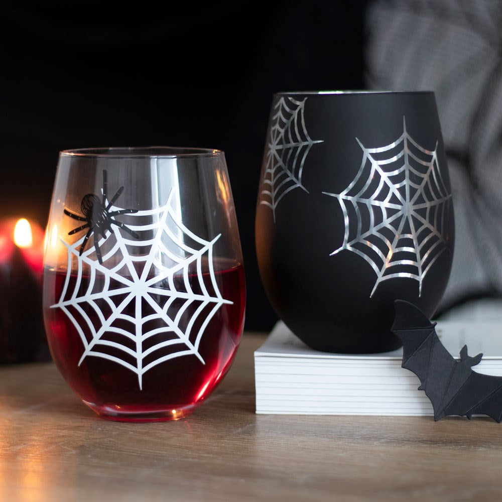 Set of two spiderweb stemless wine glasses, one clear with white spider web print and one black with silver web design, styled with red wine and candles on a wooden surface