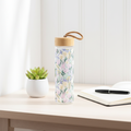 Spring floral glass water bottle with bamboo lid and loop, featuring a pastel pink and blue floral pattern, styled on a bright white surface