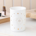 Matt white ceramic oil burner with sun moon and star cut-out design, styled on a white surface with an essential oil bottle and wooden board