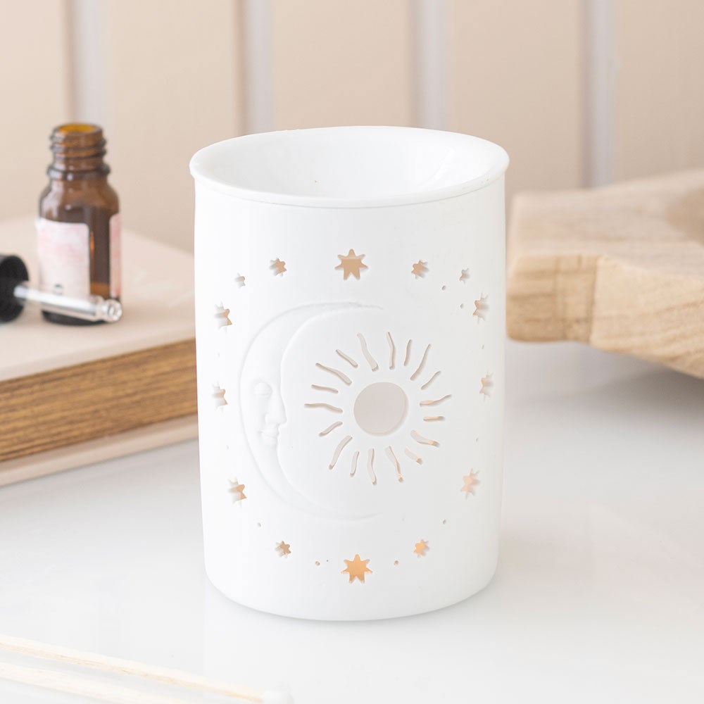 Matt white ceramic oil burner with sun moon and star cut-out design, styled on a white surface with an essential oil bottle and wooden board