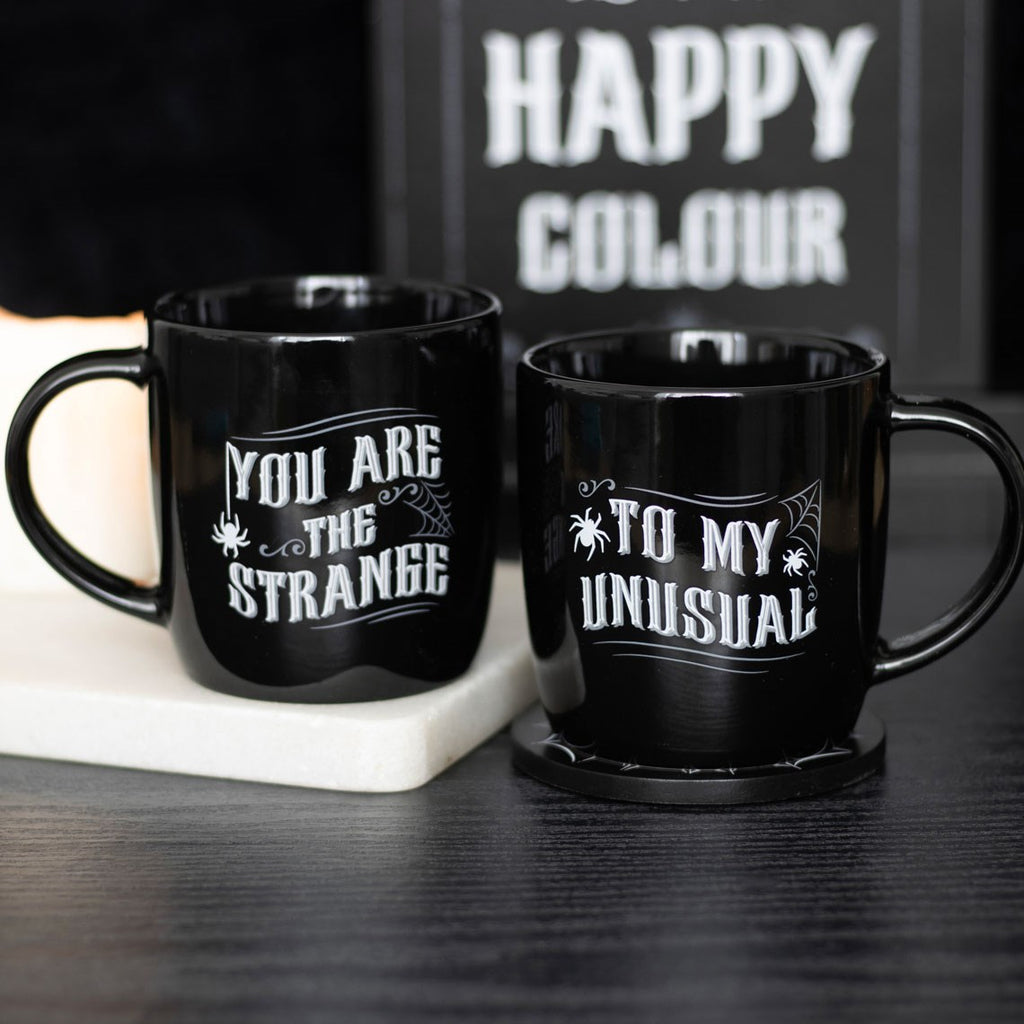 Two black ceramic mugs with white gothic lettering, one reading You Are The Strange and the other To My Unusual, with spider and cobweb details, styled on a dark surface