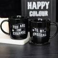 Two black ceramic mugs with white gothic lettering, one reading You Are The Strange and the other To My Unusual, with spider and cobweb details, styled on a dark surface