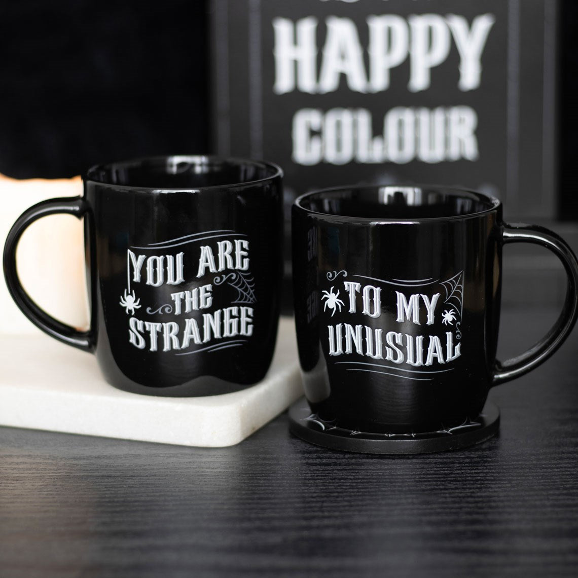 Two black ceramic mugs with white gothic lettering, one reading You Are The Strange and the other To My Unusual, with spider and cobweb details, styled on a dark surface