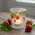 Frosted glass votive candle holder with red strawberry and green leaf design, lit tealight inside casting warm glow, styled on rustic wood with fresh strawberries