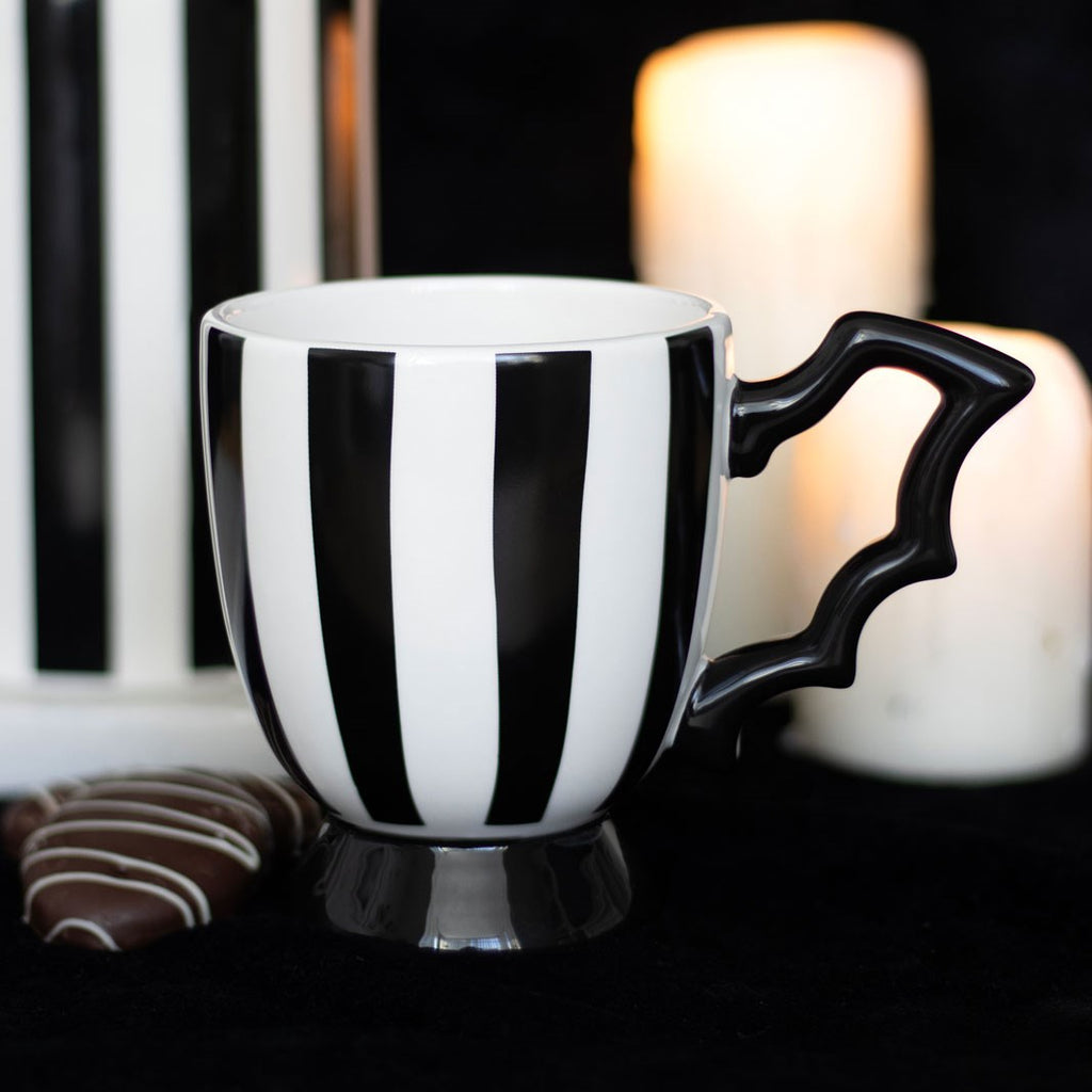 Black and white striped ceramic pedestal mug with a bat wing shaped handle, styled with candles and chocolates on a dark surface