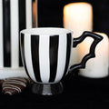 Black and white striped ceramic pedestal mug with a bat wing shaped handle, styled with candles and chocolates on a dark surface