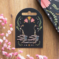 Pair of blue enamel swallow drop earrings displayed on a black folk art gift card reading "Spread Your Wings Enamel Earrings", laid on a wooden surface with pink gypsophila flowers