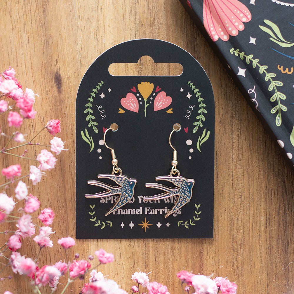 Pair of blue enamel swallow drop earrings displayed on a black folk art gift card reading "Spread Your Wings Enamel Earrings", laid on a wooden surface with pink gypsophila flowers