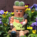 Terracotta man plant pot with a painted smiley face, red nose, green dungarees and gardening gloves, planted with a cactus and surrounded by colourful pansy flowers in a garden setting