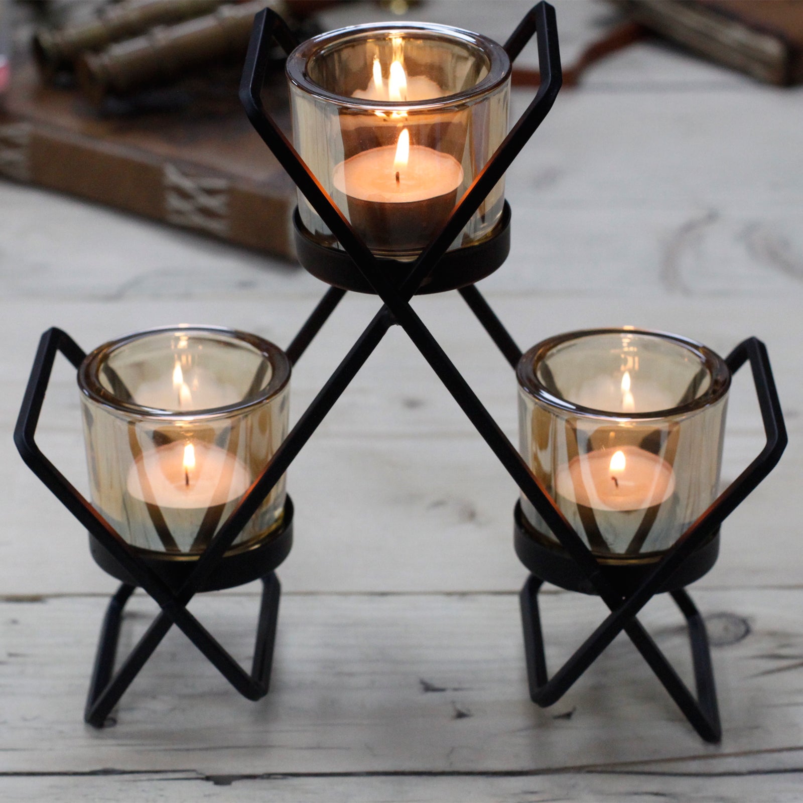 Black iron triangle centrepiece candle holder with three amber glass votive cups at varying heights, on a rustic wooden surface