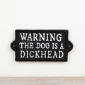 Cast iron sign reading “Warning The Dog Is A Dickhead” with rustic vintage-style humorous design.