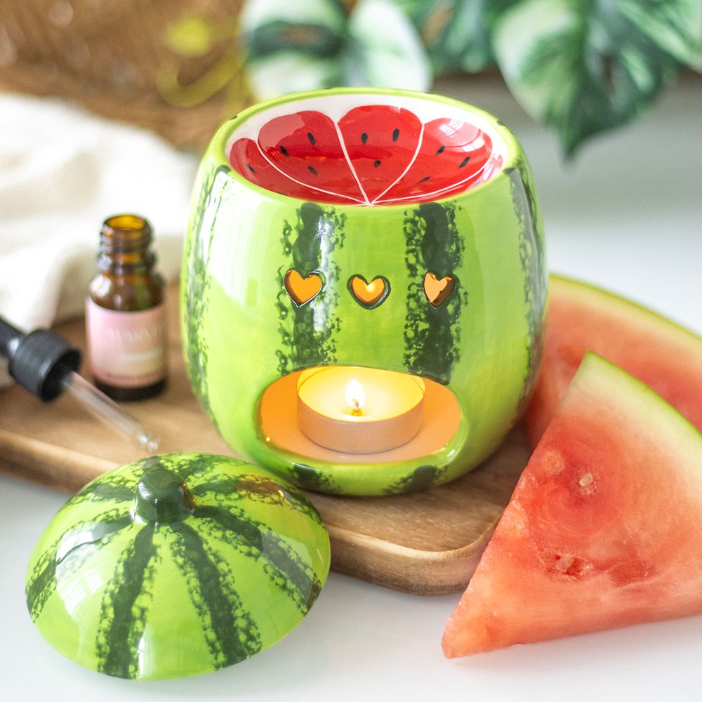 Watermelon shaped ceramic oil burner with heart cut-outs and lit tealight, styled on a wooden board with fresh watermelon slices and an essential oil bottle