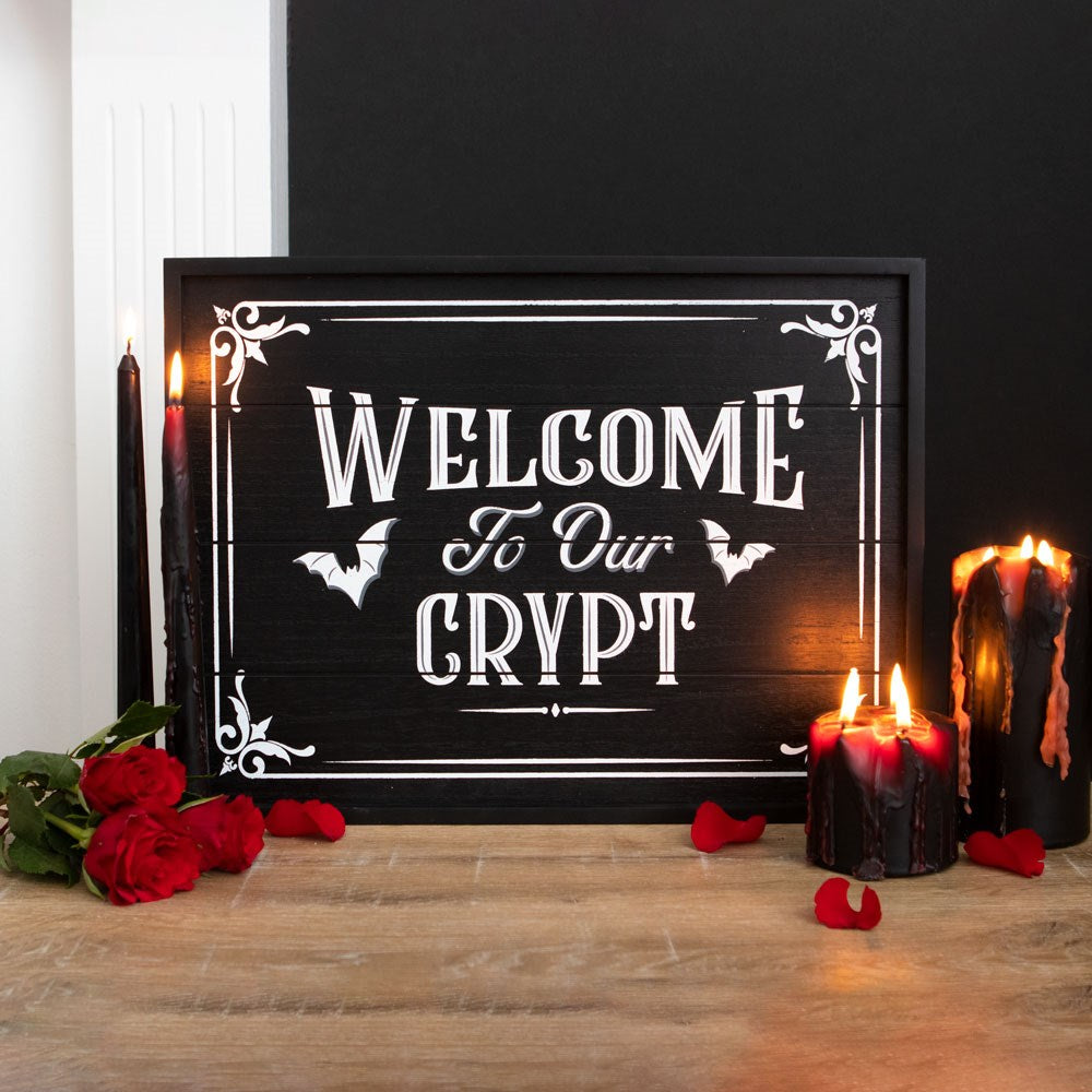 Black framed wooden wall sign with white gothic lettering reading Welcome To Our Crypt with bat and decorative corner details, styled with black and red candles and red roses